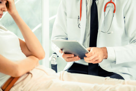 Doctor holding a tablet sitting beside a patient laying on a bed
