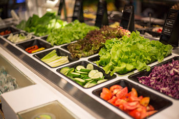 A white salad bar with small black container inserts full of lettuce, tomatoes, cucumbers, bell peppers, onions, and celery