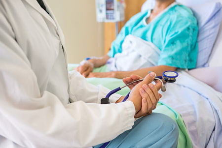 Doctor holding a stethoscope sitting beside a patient on the side of the patient\'s bed