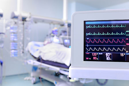 Vital signs monitor in the foreground with a hospital bed and equipment in the backgound